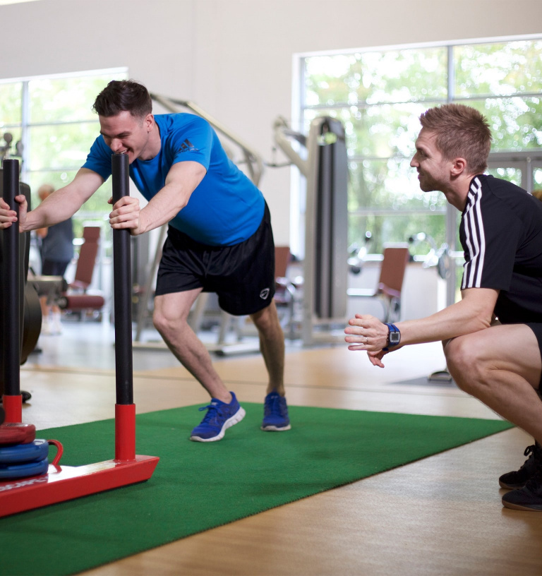 Male PT assisting male client and encouraging him on the sled push.